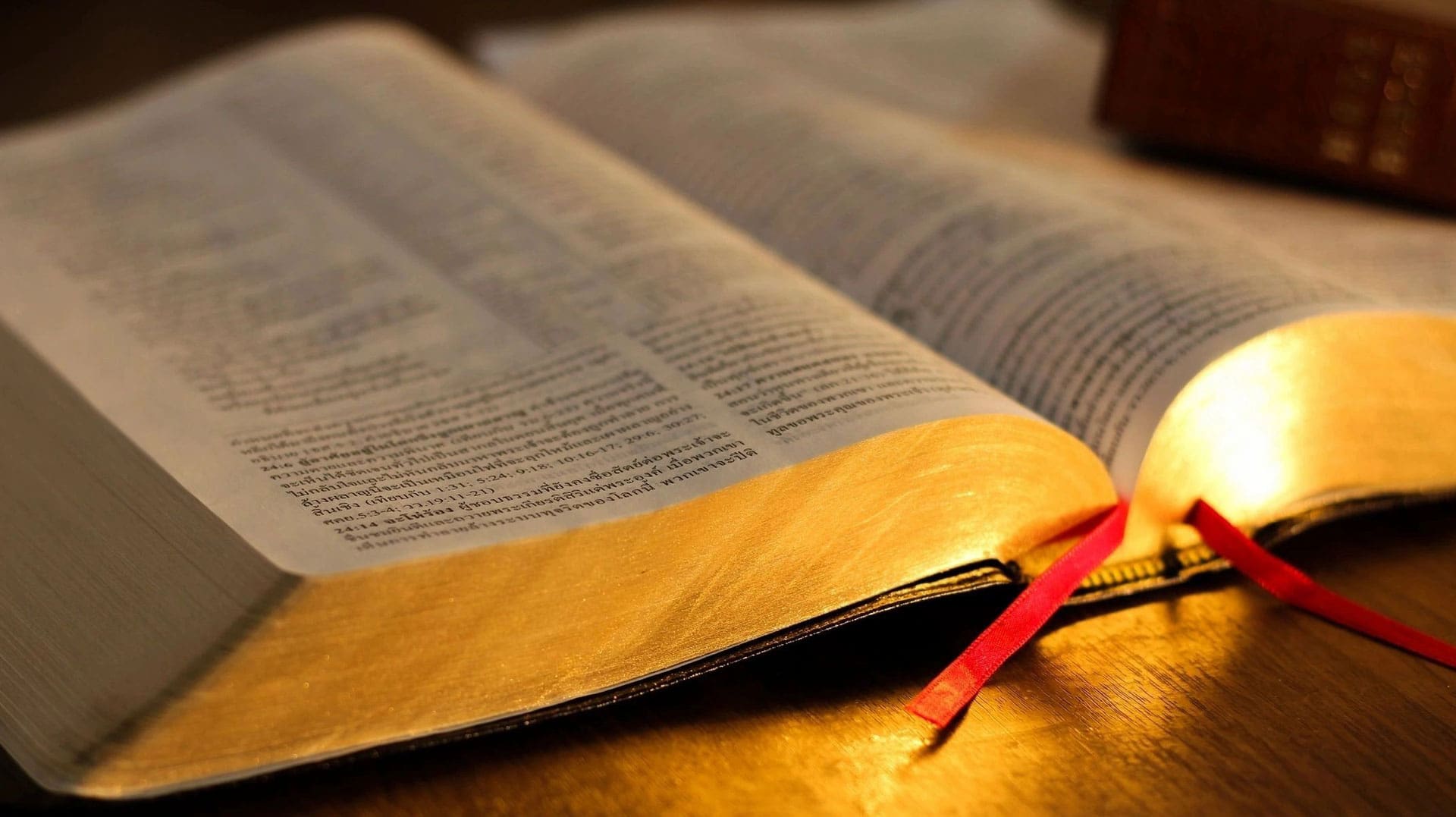 Open Bible with gilded page edges and a red ribbon marker, softly lit on a wooden table.